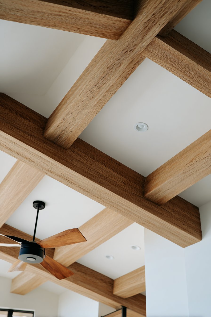 Light oak ceiling beams on a white ceiling with recessed lights and a black fan with wooden blades