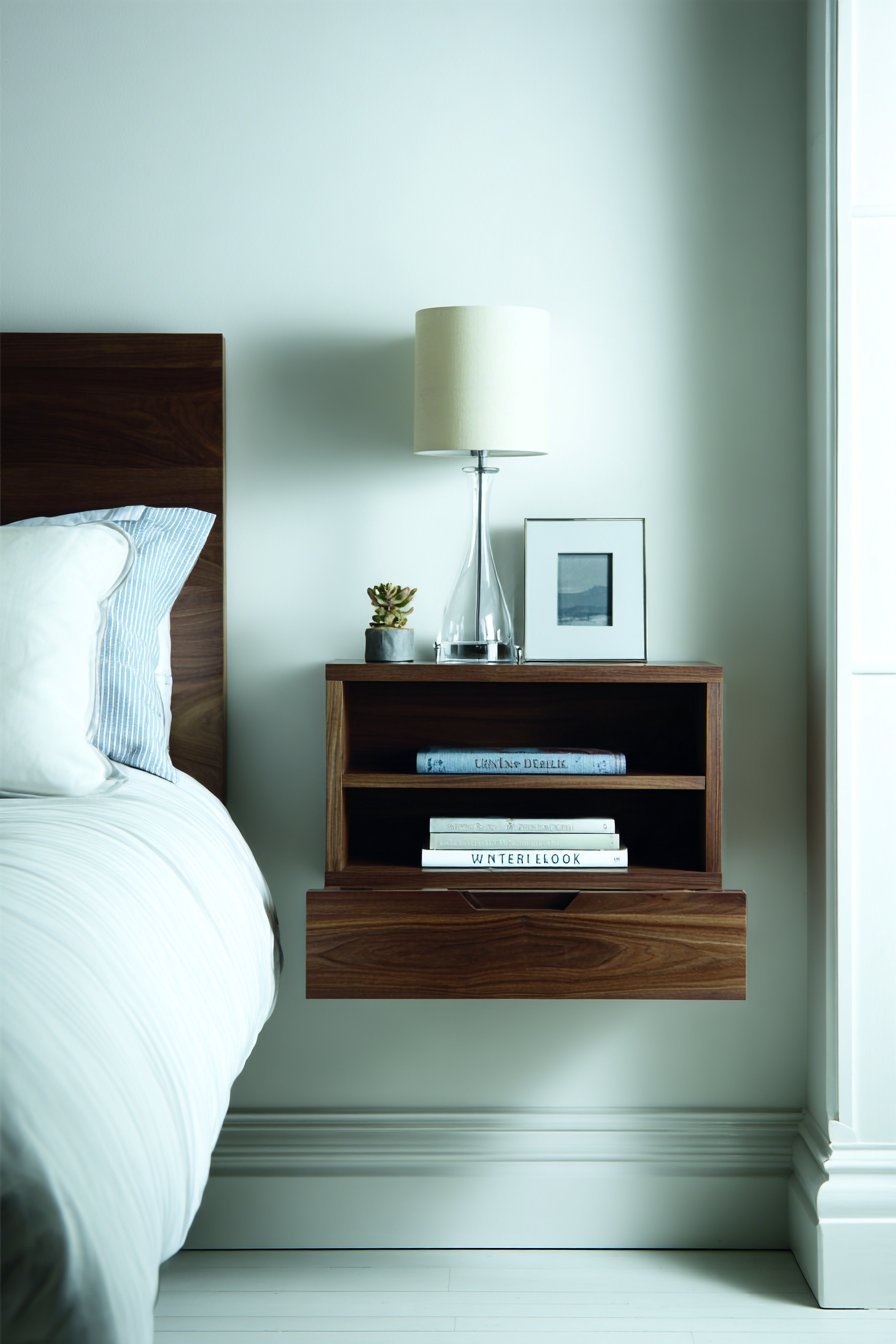 Walnut floating nightstand with one shallow drawer and open shelf holding books and a table lamp on a white wall