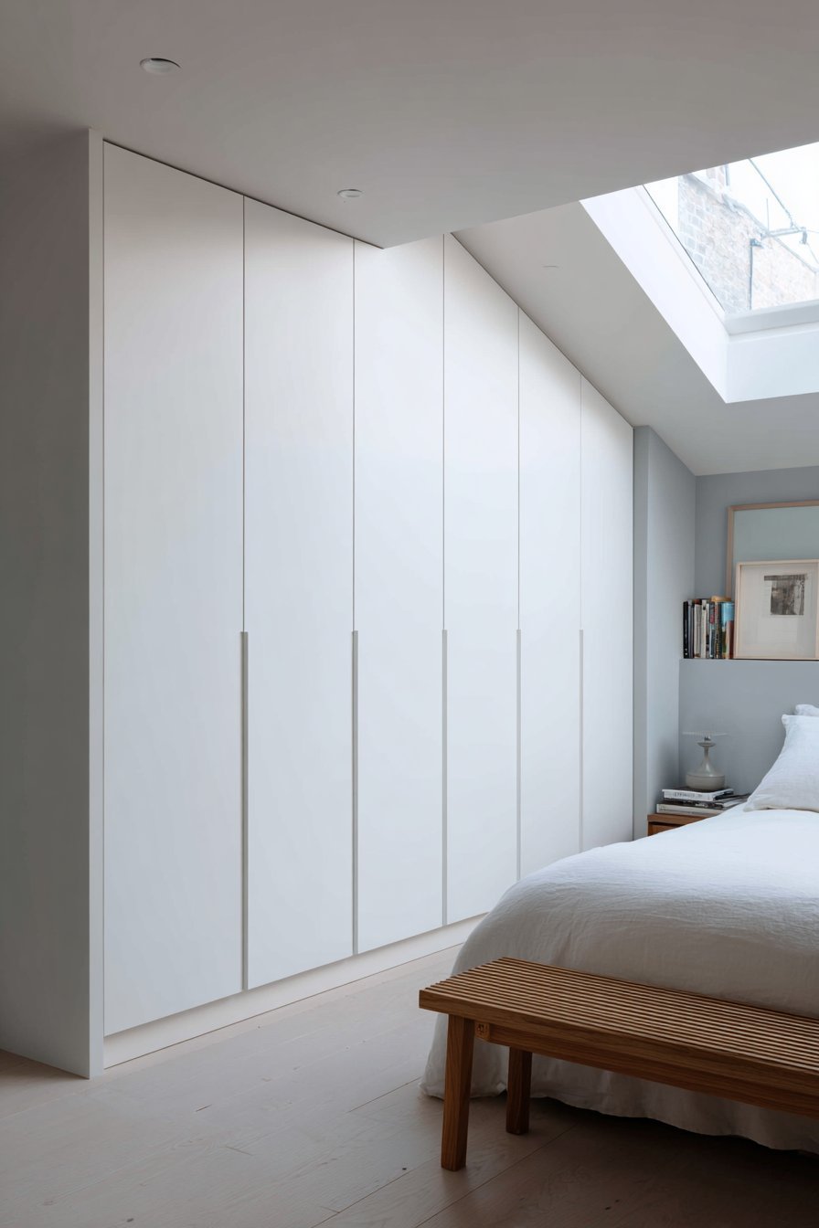 Handleless white matte wardrobes spanning one wall beside an oak bench and bed under a skylight