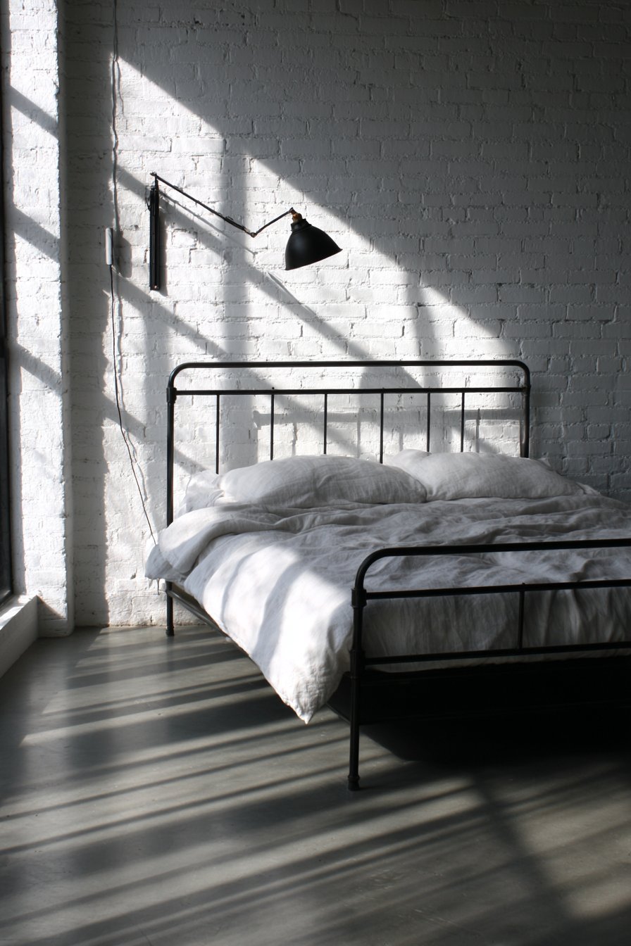 Matte black metal bed with white linen sheets, wall-mounted reading lamp and polished concrete floor beside white brick