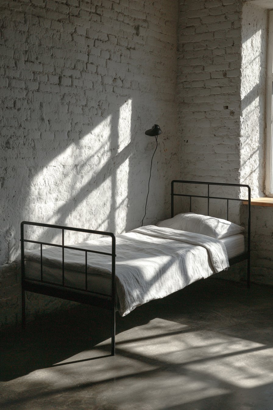 Matte black metal bed with white linen sheets, wall-mounted reading lamp and polished concrete floor beside white brick