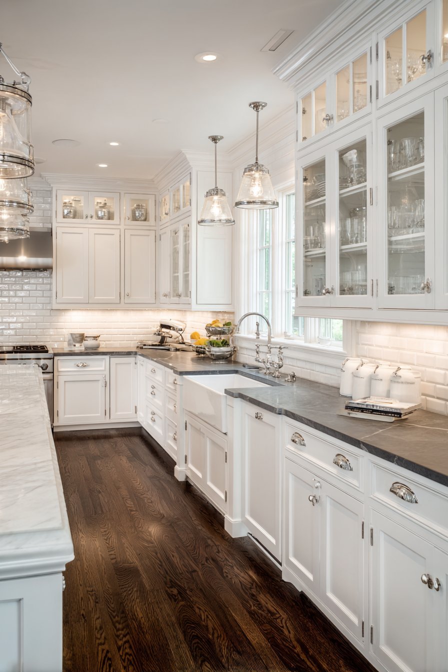 White kitchen with glass-front uppers, soapstone counters, apron sink, schoolhouse pendants and dark hardwood floor