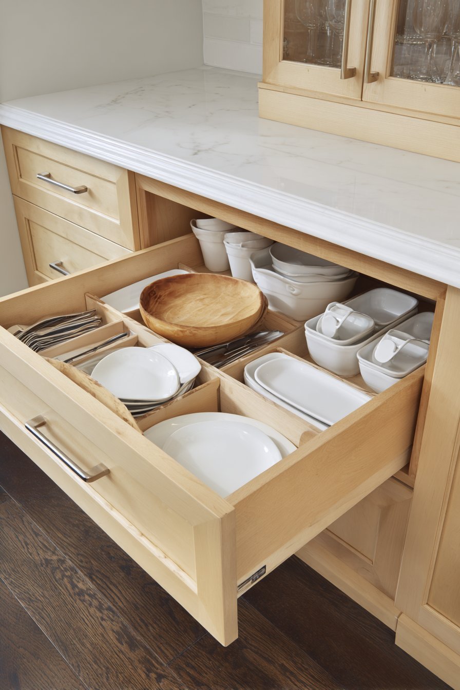 Open drawer with custom dividers beside glass-front oak upper cabinets and white solid surface counters