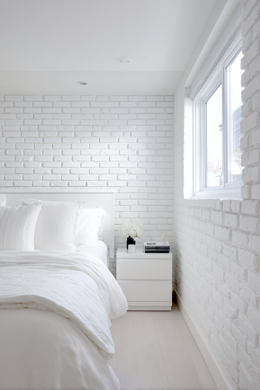 All-white small bedroom with painted brick wall, layered linen bedding and light ash wood floor