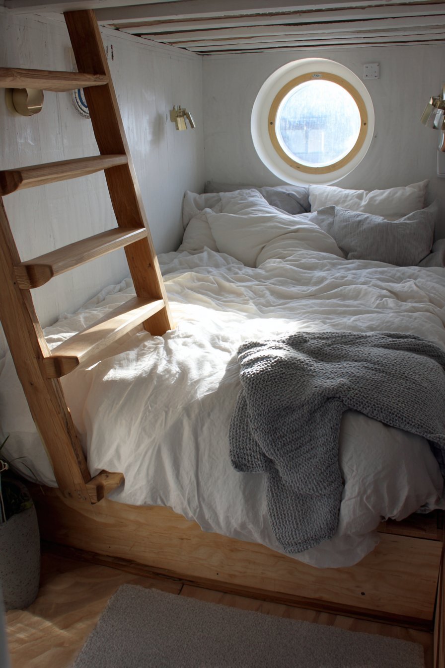 Tiny house loft bed reached by a maple ladder, with white sheets, grey knit blanket and a porthole window