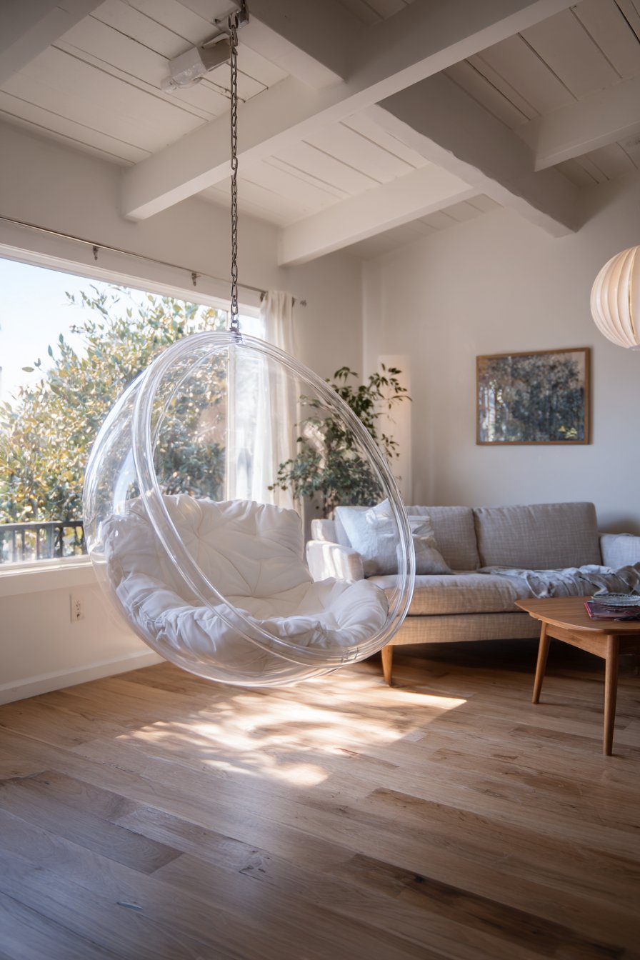 Hanging clear acrylic bubble chair with white cushions beside a window and warm grey linen sofa