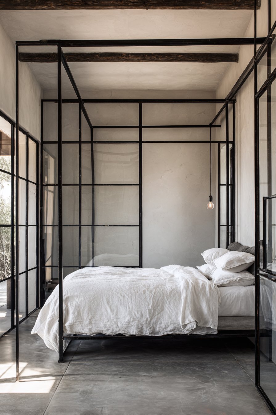 Rusted raw steel tube bed frame with white bedding, polished concrete floor, Edison bulb and steel-framed windows