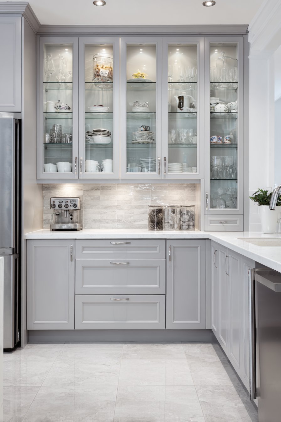 Glass-front upper cabinets with interior lighting above medium grey shaker lowers and white quartz