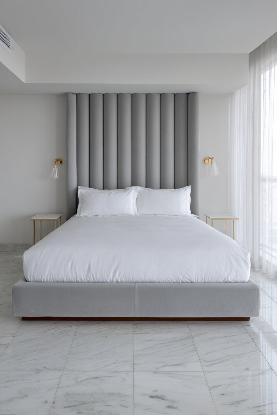 Slim ceiling-height grey velvet channel headboard, white hotel bedding, brass swing-arm lamps and white marble floor