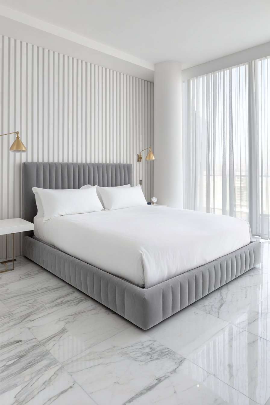 Slim ceiling-height grey velvet channel headboard, white hotel bedding, brass swing-arm lamps and white marble floor