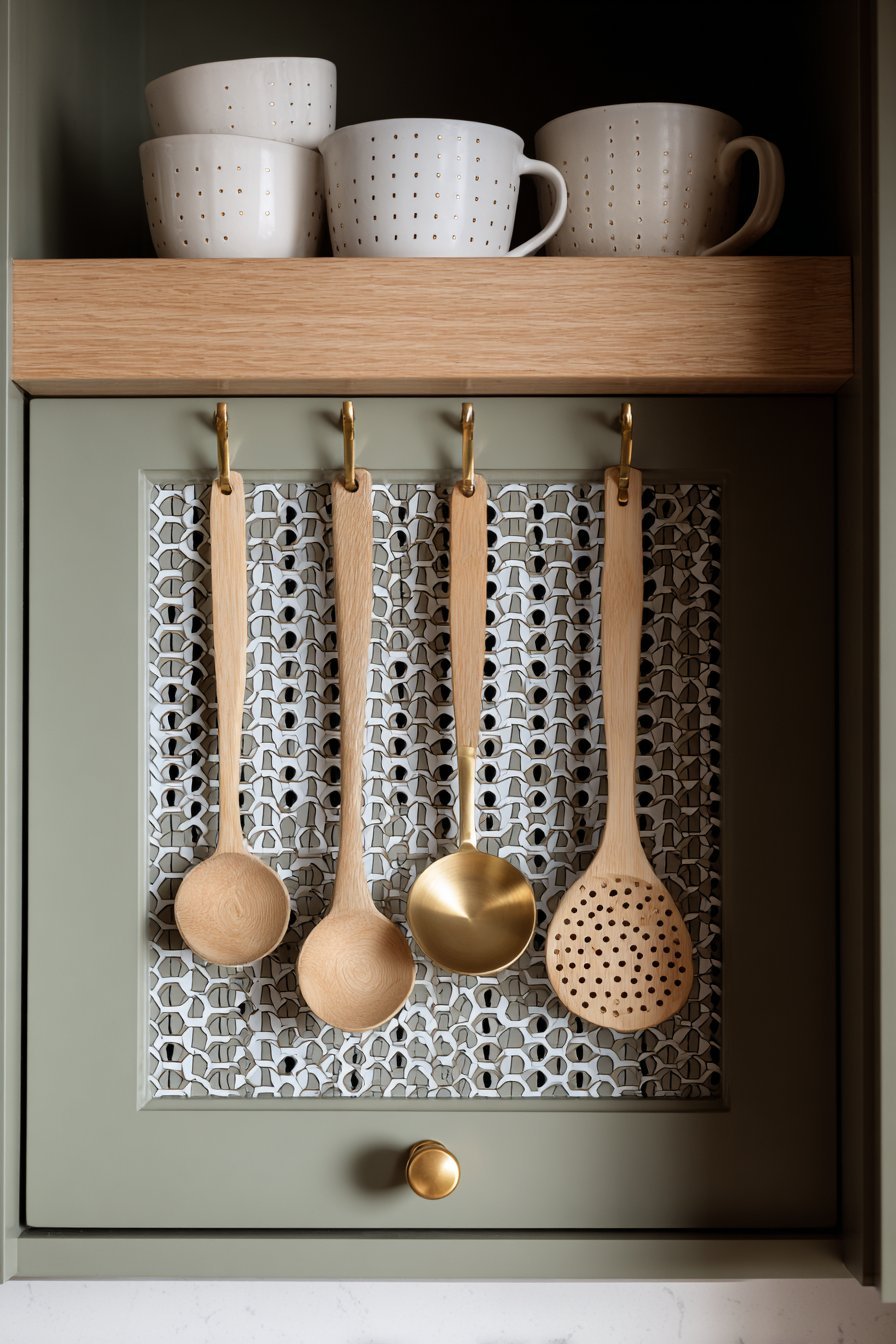 Pegboard inside a sage green cabinet door holding measuring cups and spoons beside oak floating shelves