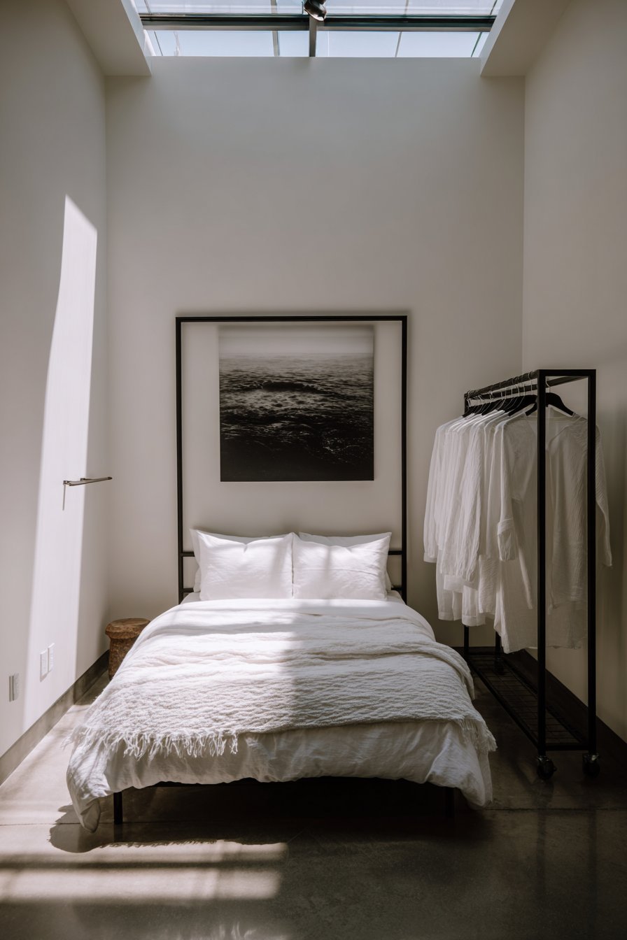 Matte black metal bed, rolling black clothing rack, large black and white print and skylight on grey concrete