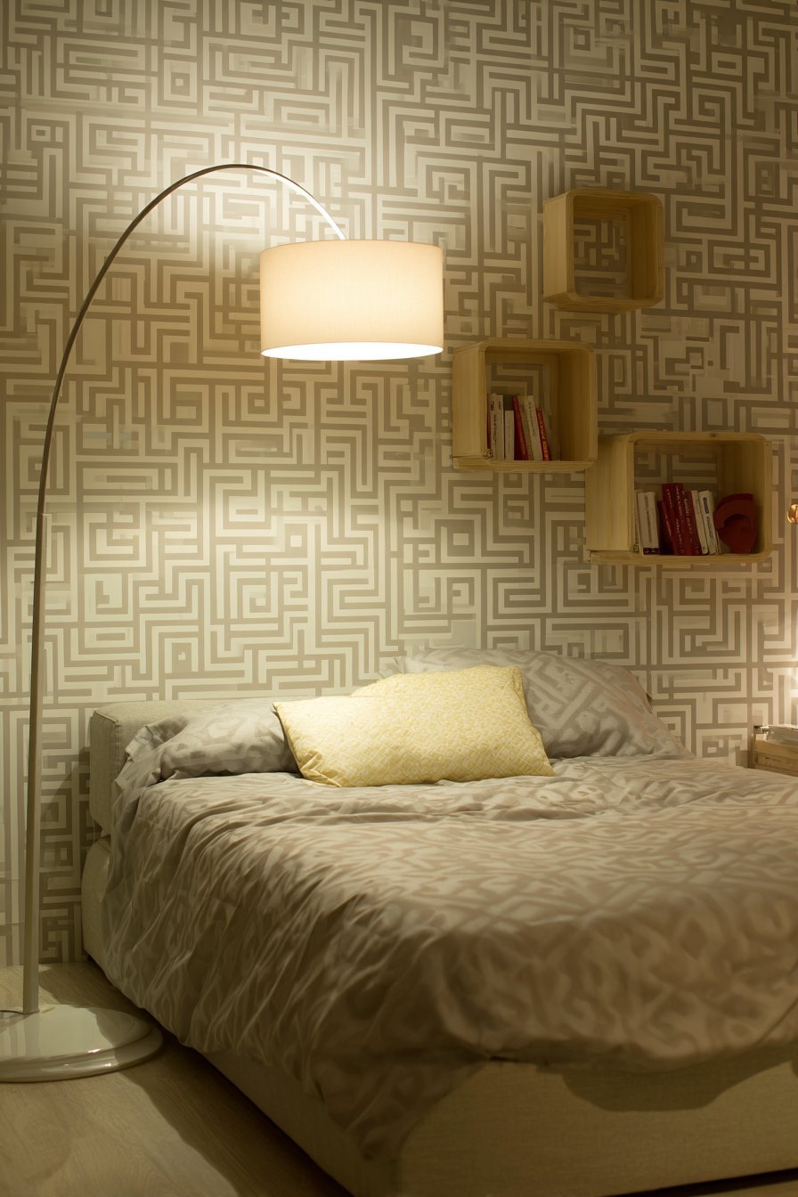 Grey and cream geometric wallpaper behind a taupe upholstered bed with floating shelves and arc floor lamp